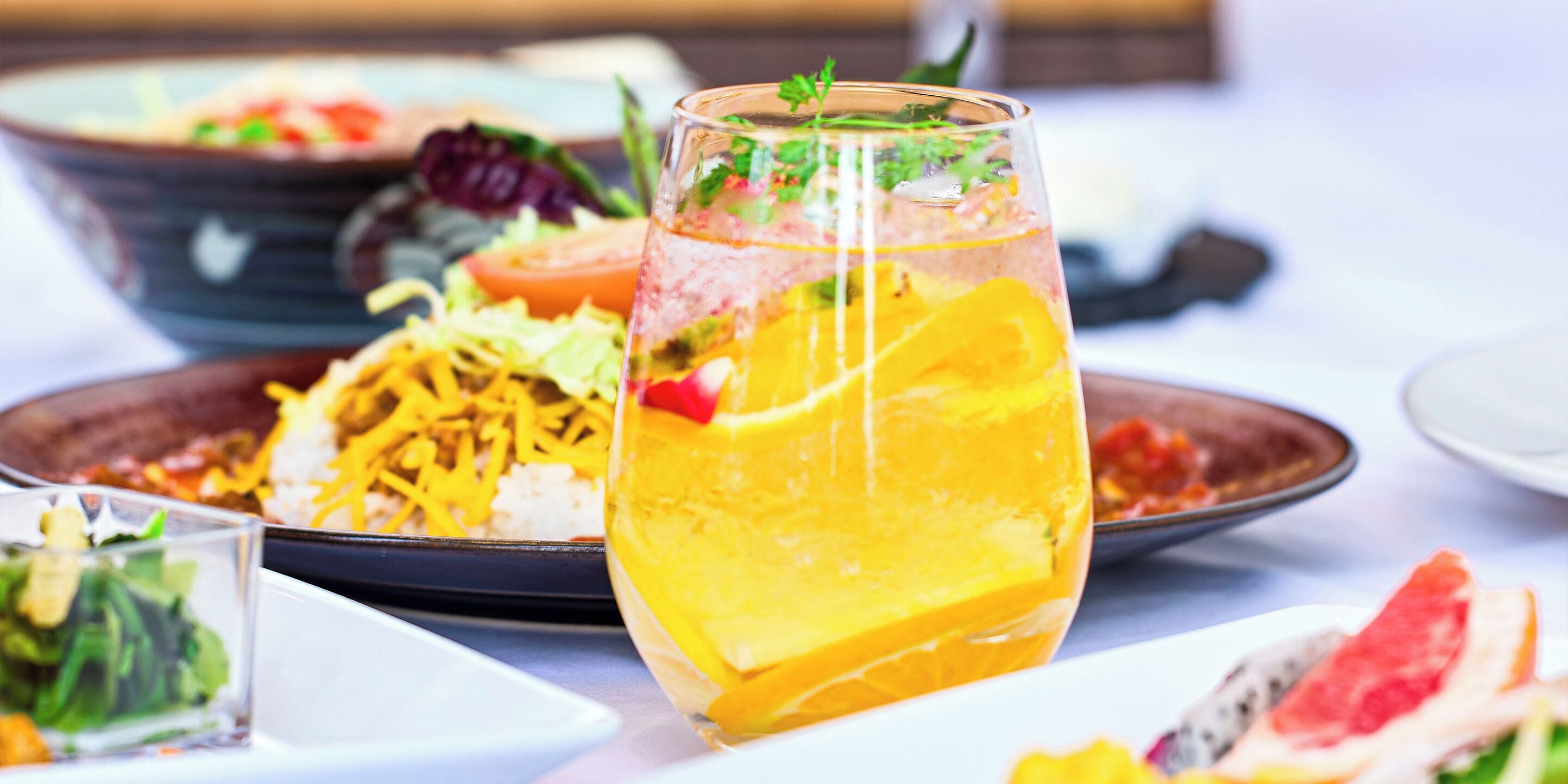 Een glas sinaasappelsap met een partje sinaasappel en munt, omringd door diverse gerechten, waaronder een salade en een schaal eten.