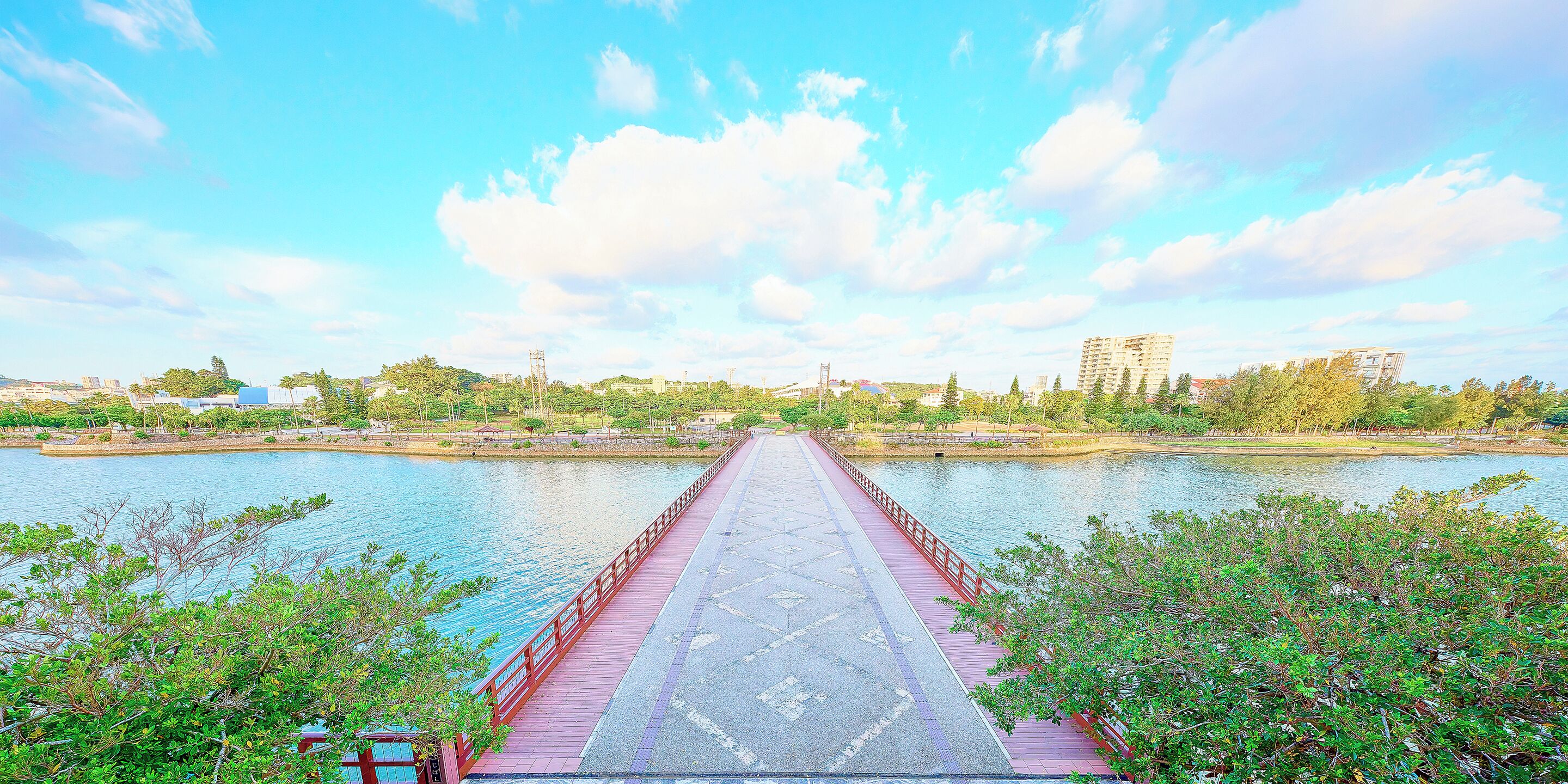A long red bridge over a calm river with buildings and trees in the background.