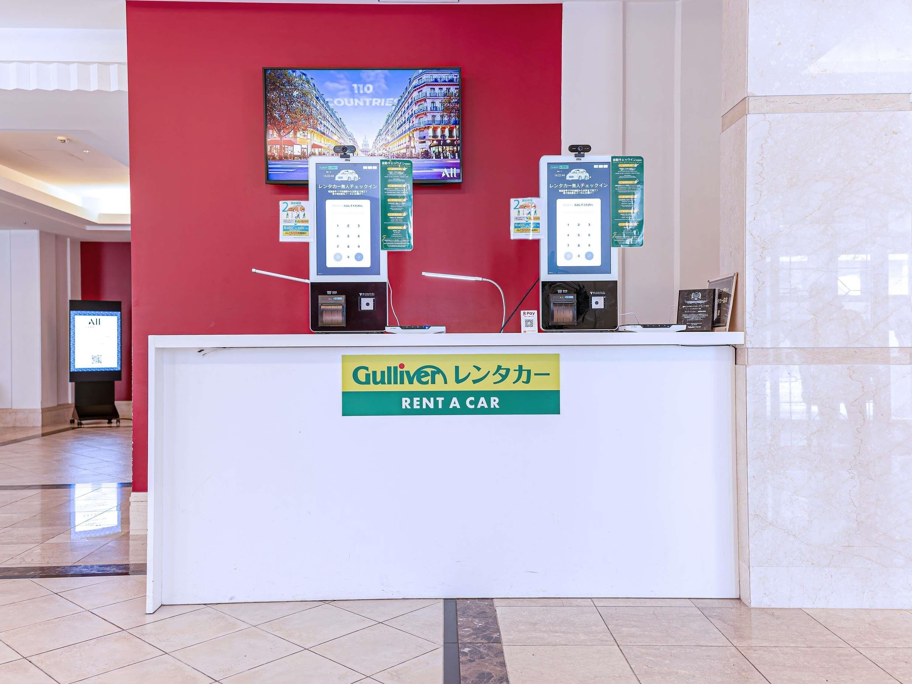 A rental car counter with a digital display, a sign for Gulliver Rent a Car, and a large screen in the background.