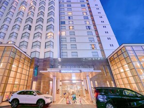 A hotel entrance with a modern design, featuring a prominent sign, a glass facade, and a parking area with cars.