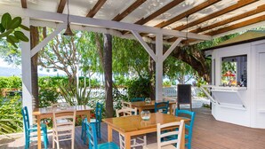 An outdoor patio with wooden tables and chairs, surrounded by greenery and a view of the sea.