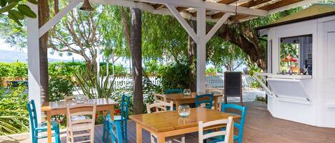 An outdoor patio with wooden tables and chairs, surrounded by greenery and a view of the sea.