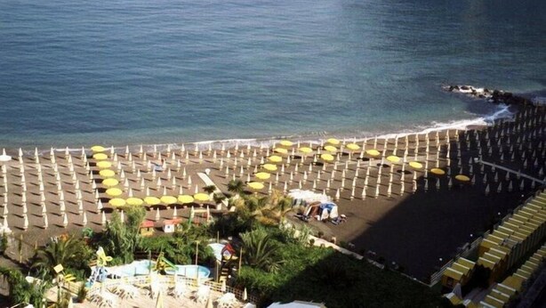 A beach with rows of umbrellas and sun loungers, a pool area, and a view of the ocean.