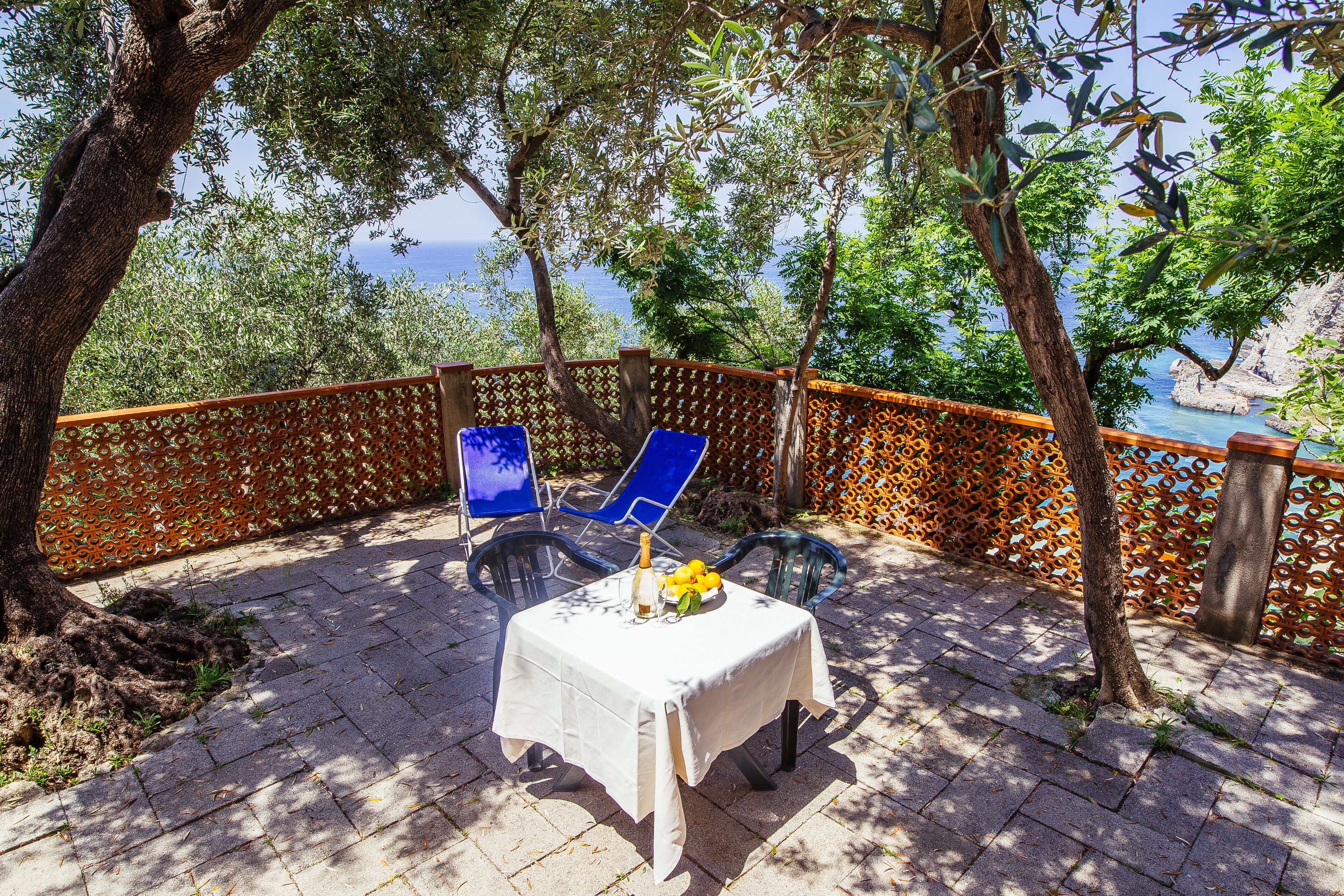 A table with a white tablecloth, two blue chairs, and a bottle of champagne on a paved patio with a view of the sea.