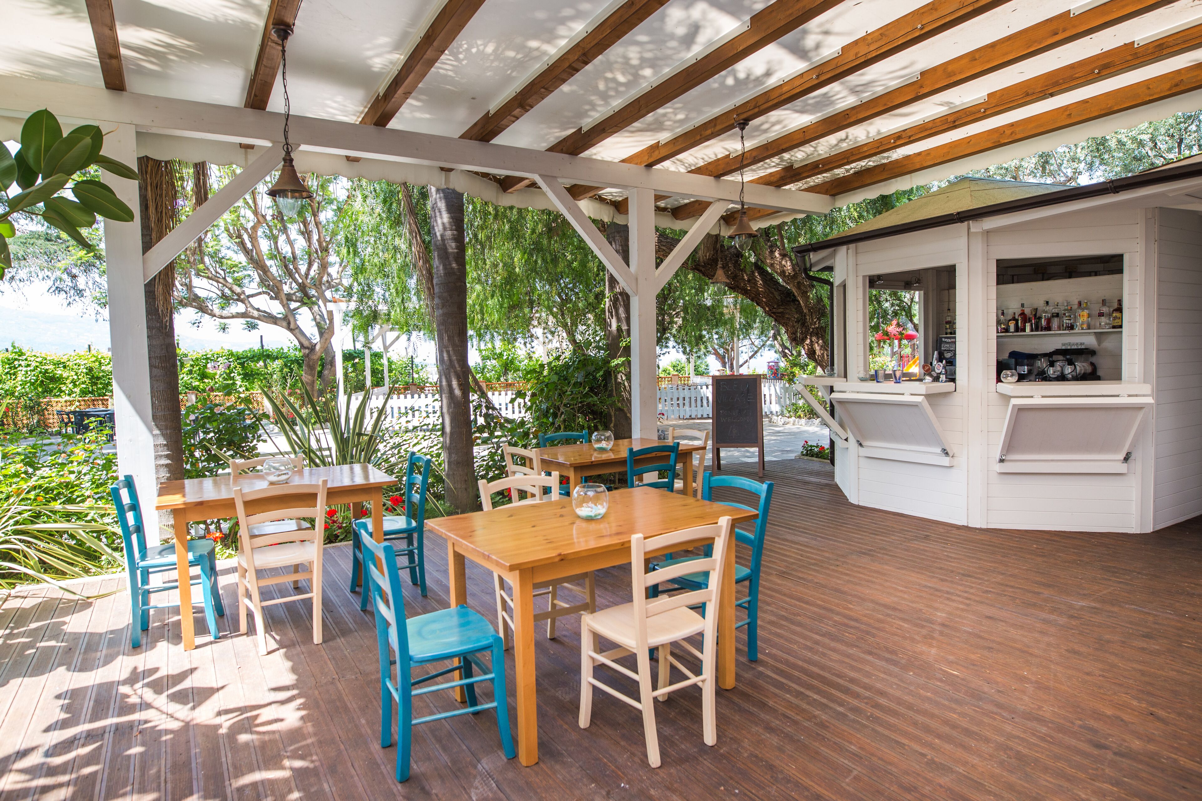 Een terras buiten met houten tafels en stoelen, een bar met flessen op de planken, en uitzicht op groen en bomen.