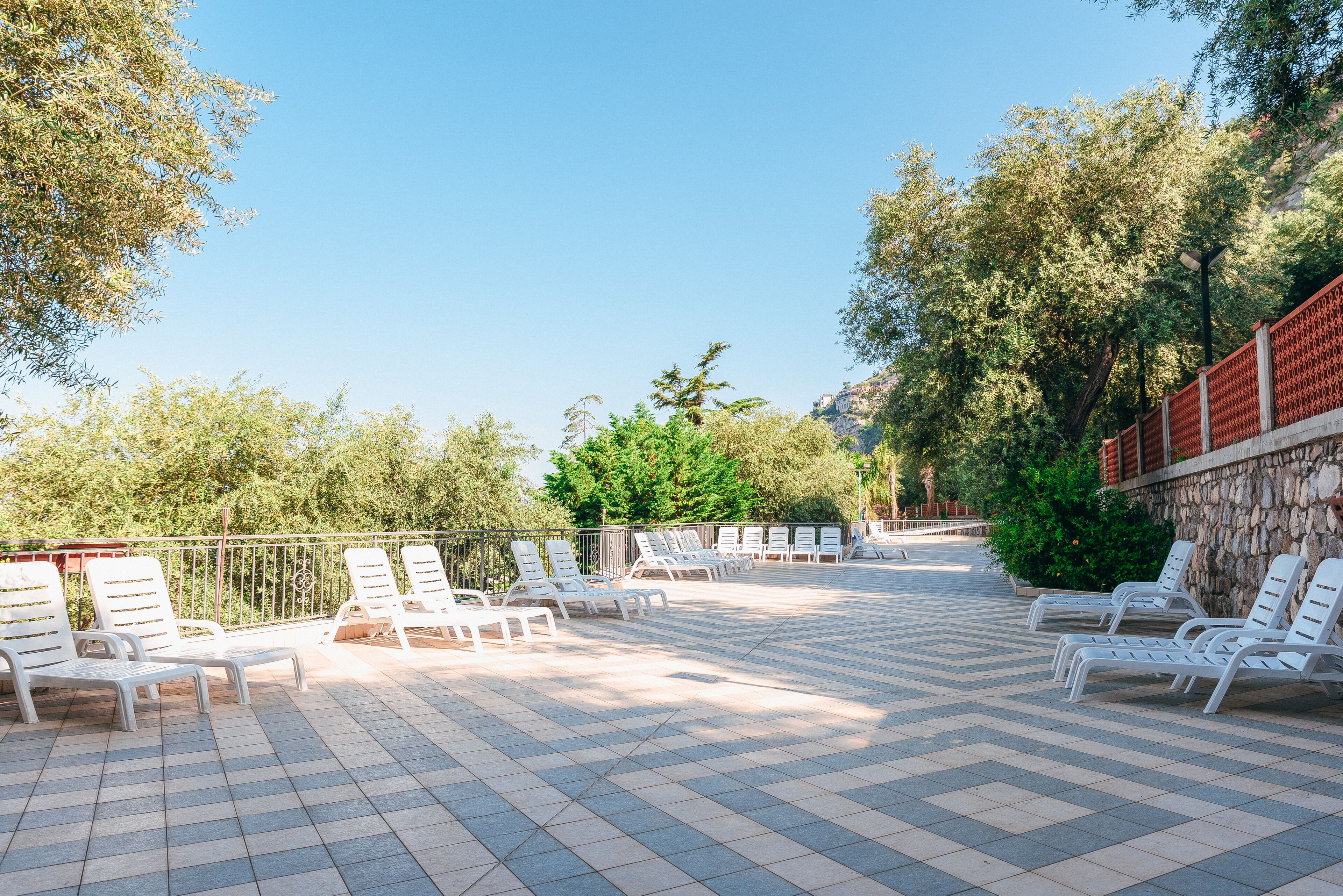 Ein sonniger Außenbereich mit Sitzgelegenheiten, weiße Loungestühle sind in Reihen angeordnet, eine Steinmauer an einer Seite und Bäume im Hintergrund.