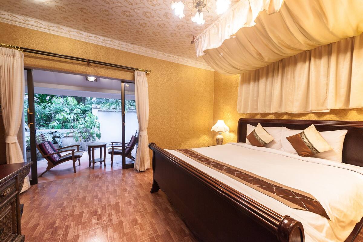 A hotel room with a large bed, a sitting area with chairs, a view through a glass door, and a chandelier.