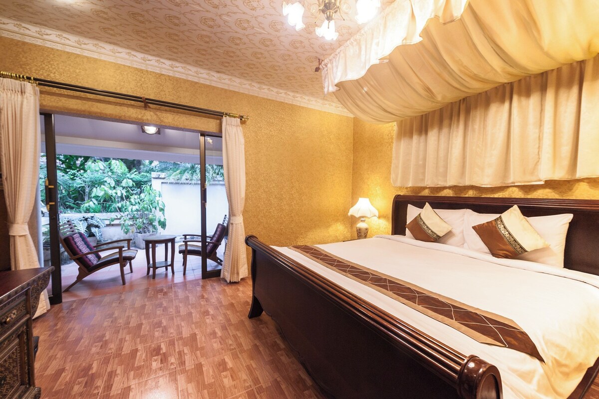Quarto de hotel com uma cama grande, um espaço de estar com cadeiras, vista através de uma porta de vidro e um candelabro.
