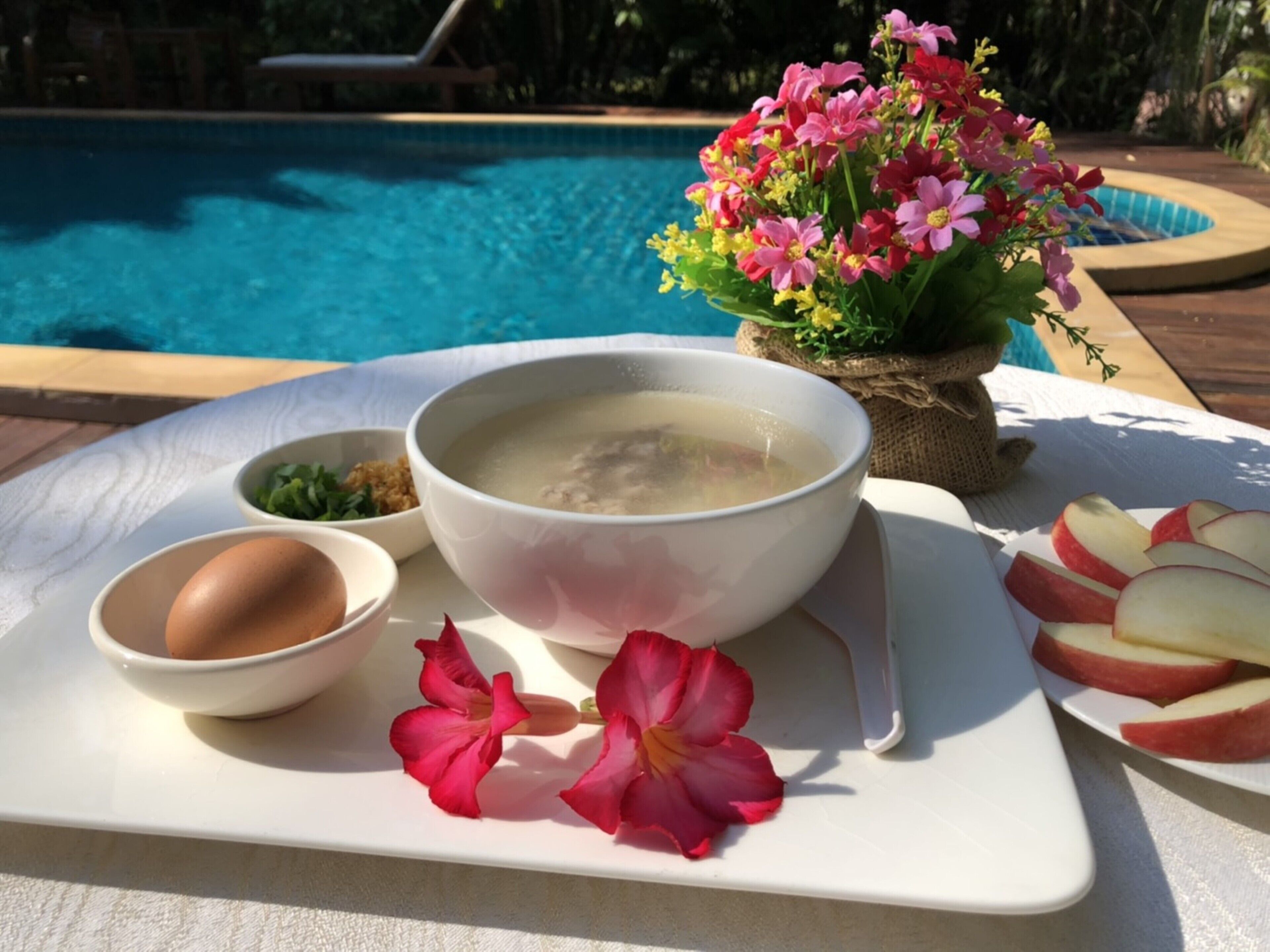 Una mesa de comedor con un tazón de sopa, un huevo cocido, rodajas de manzana y una flor sobre una bandeja blanca, cerca de una piscina.