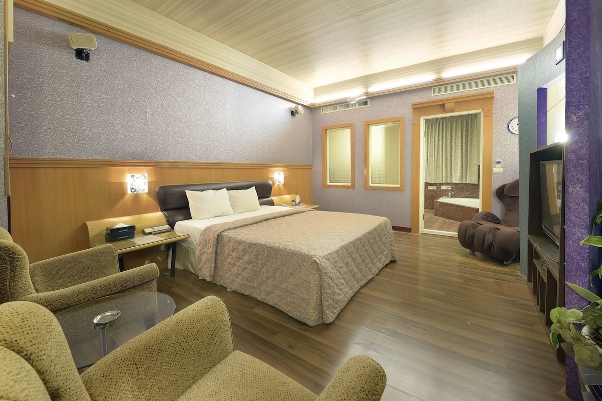 Una camera d'albergo con un letto, un'area salotto con un tavolo, una televisione e un bagno visibile attraverso una porta aperta.