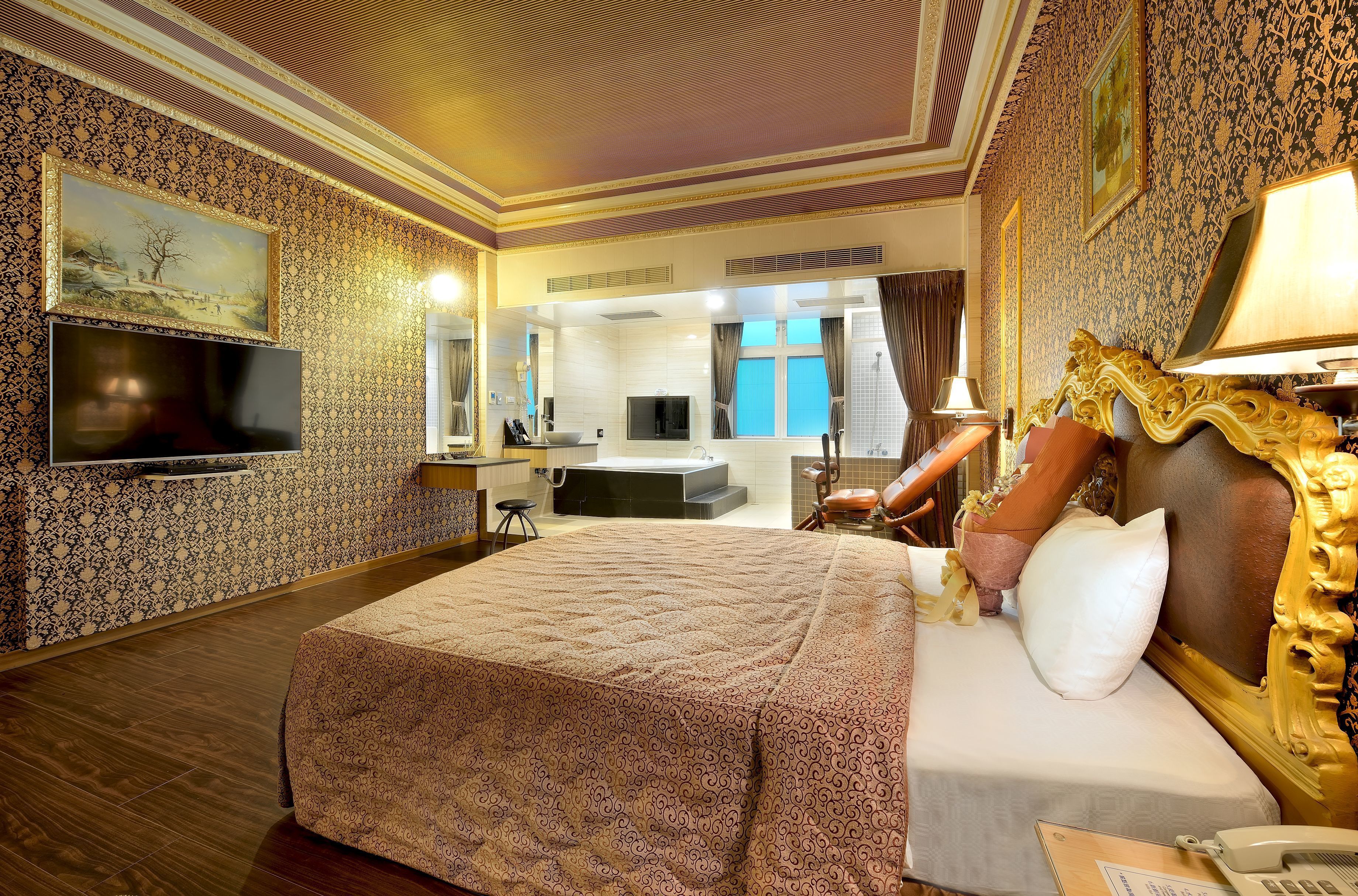 A hotel room with a large bed, a TV mounted on the wall, a bathroom visible through an open door, and a mirror above the bed.