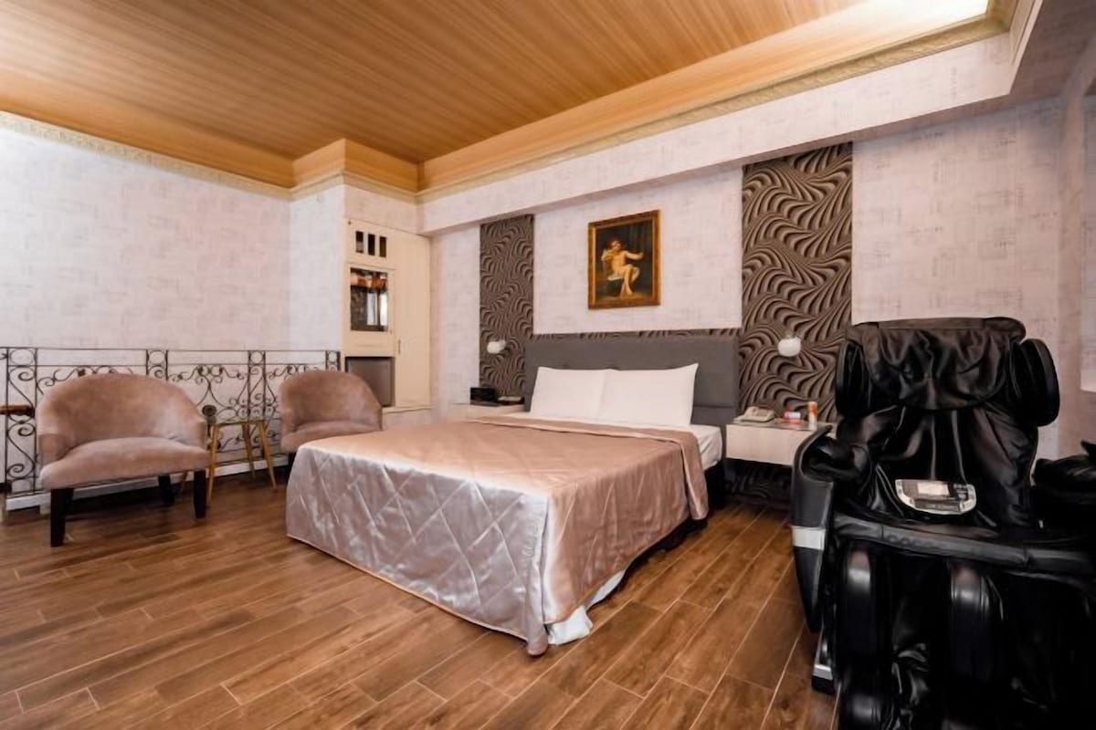 A bedroom with a bed, two armchairs, a massage chair, a painting, and a patterned wall.