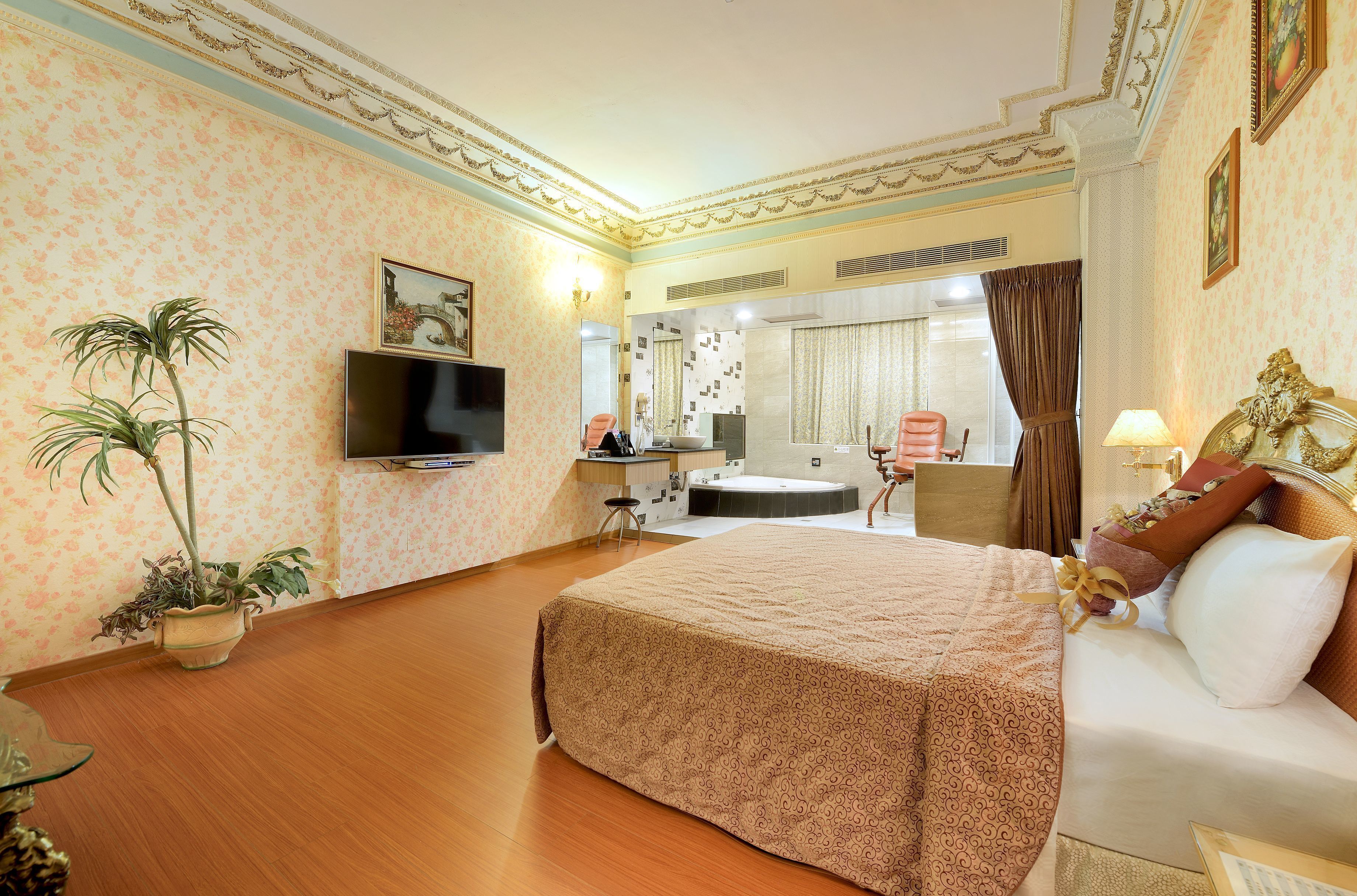 A hotel room with a large bed, a TV, a desk with a chair, a bathroom with a bathtub, and a potted plant.