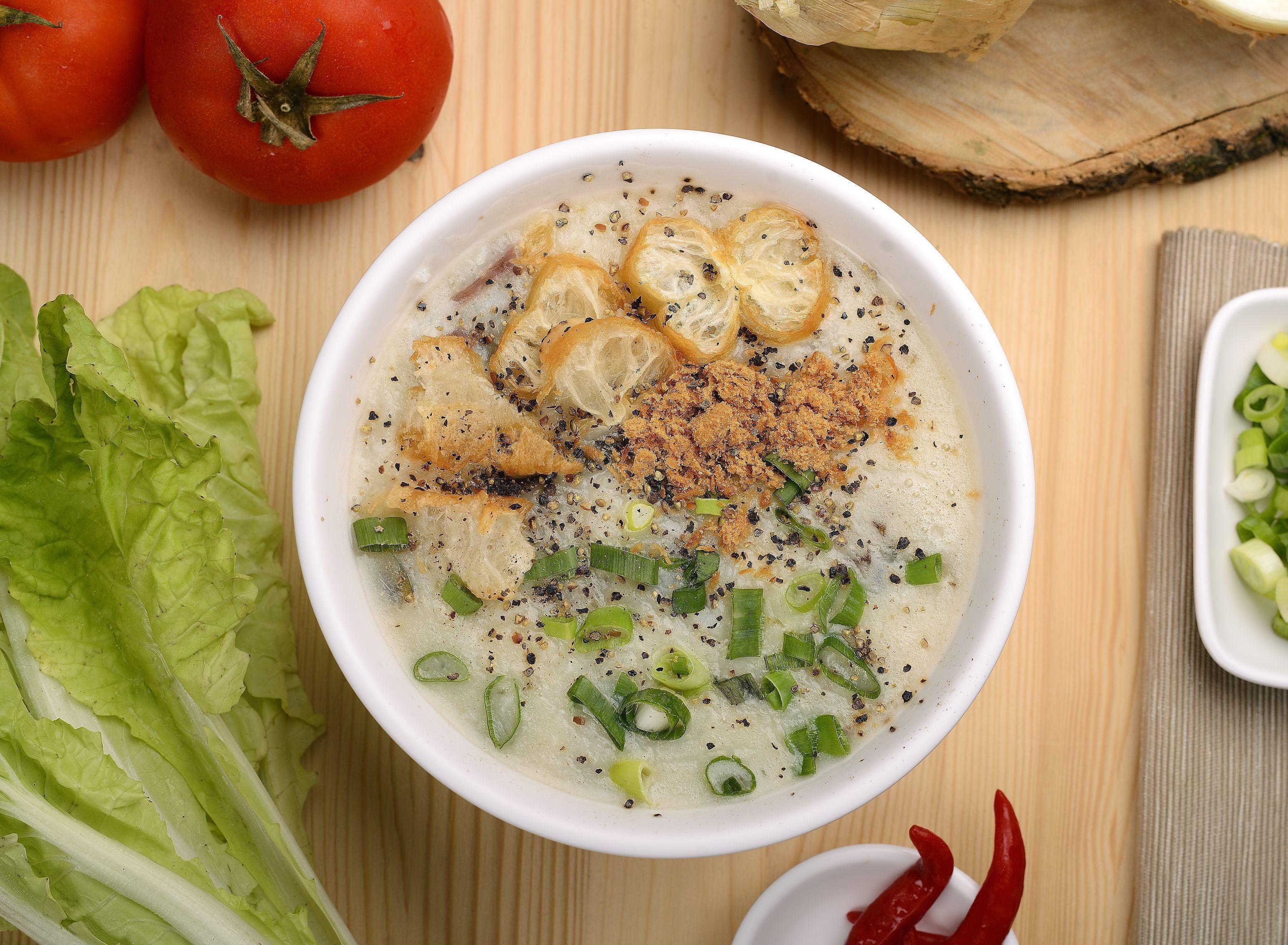 A bowl of soup with toppings, surrounded by vegetables and a wooden background.