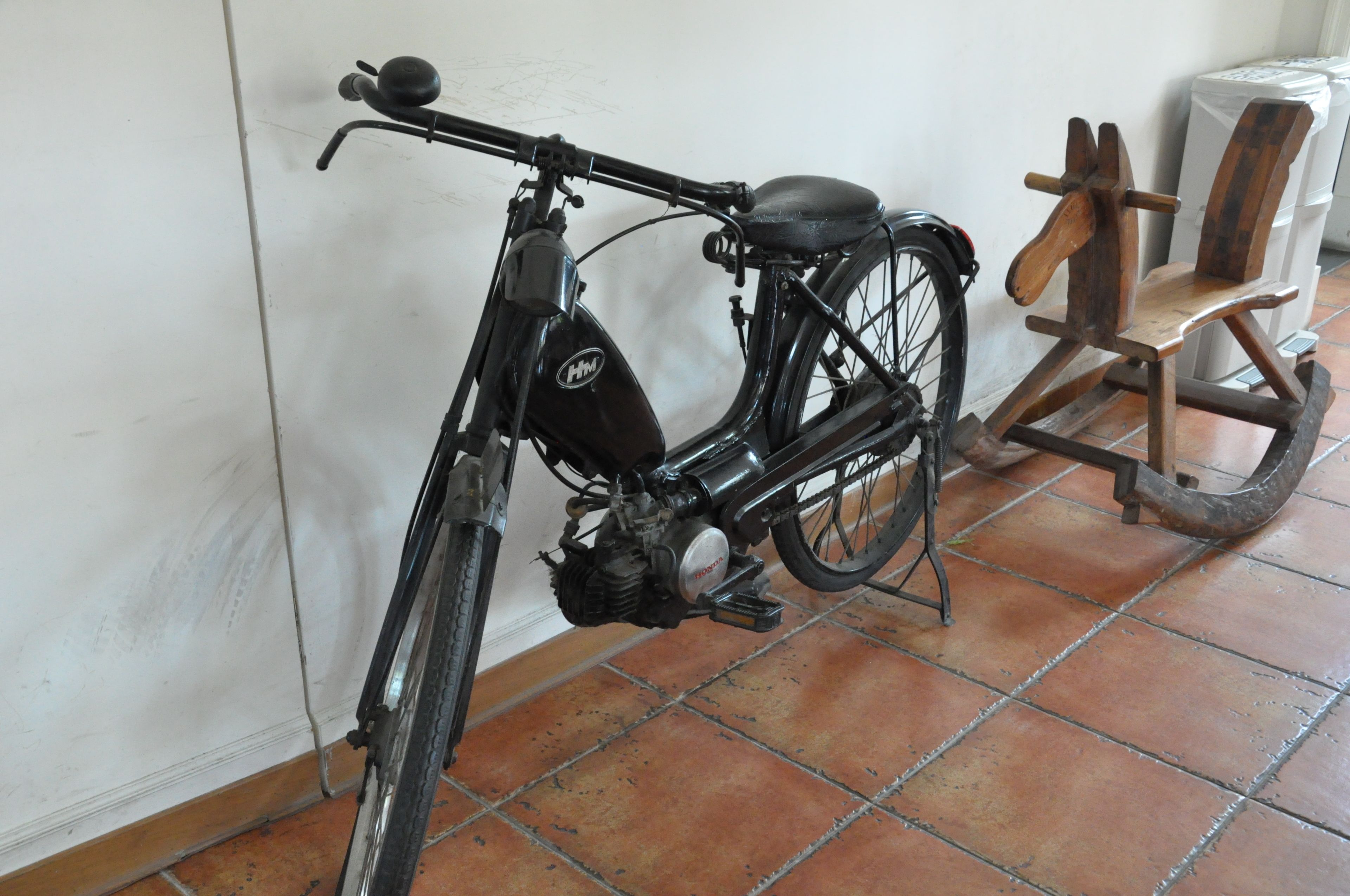 A vintage bicycle with a wooden seat and a metal frame, leaning against a wall in a room with tiled flooring.