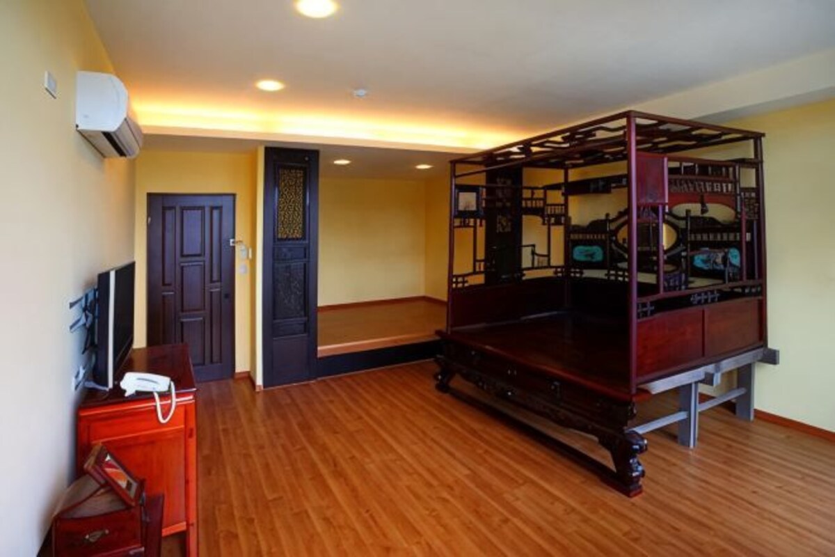 A room with a wooden floor, a bed with a metal frame, a desk with a telephone, and a door with a decorative panel.