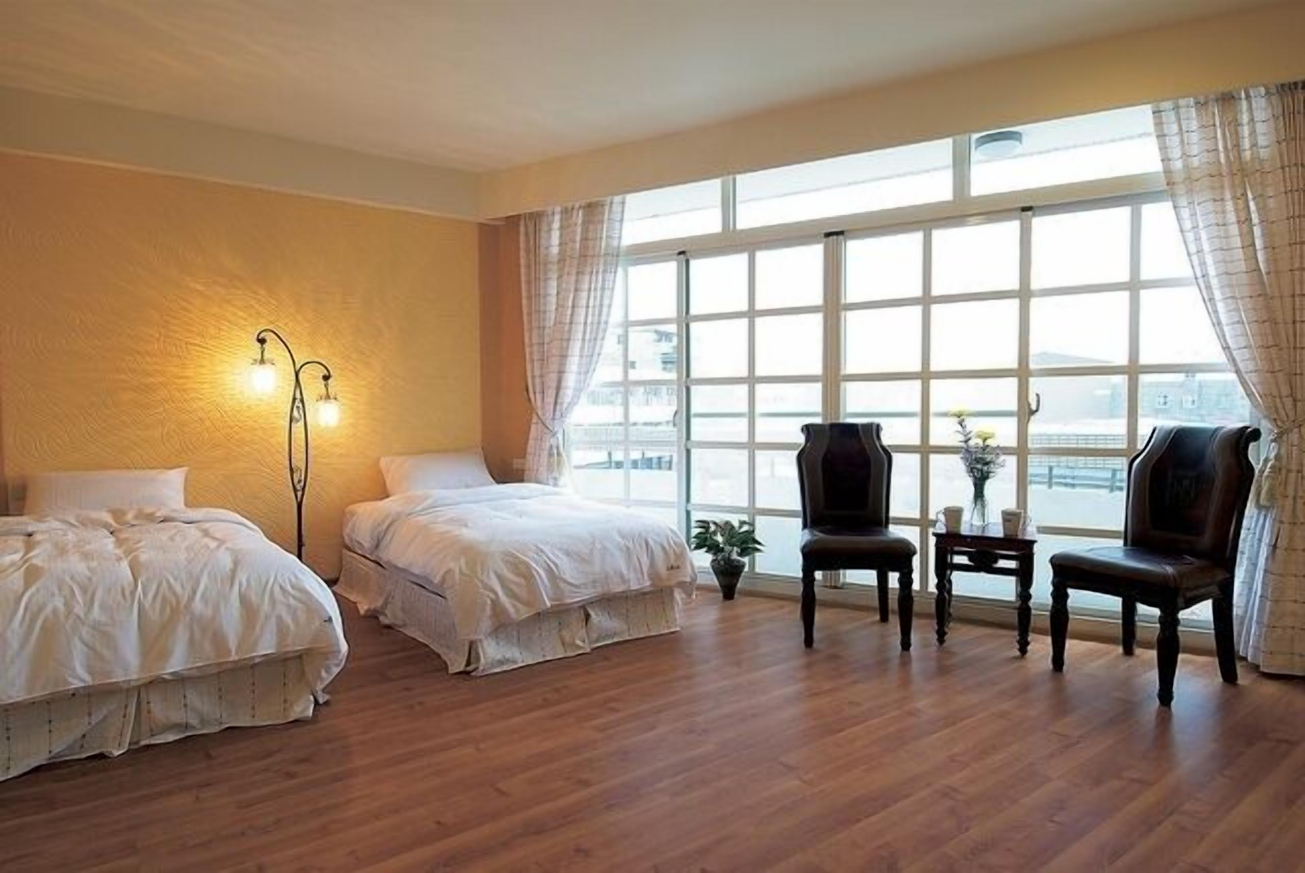 Habitación de hotel con dos camas, un escritorio con sillas, un ventanal grande y una lámpara de pie.