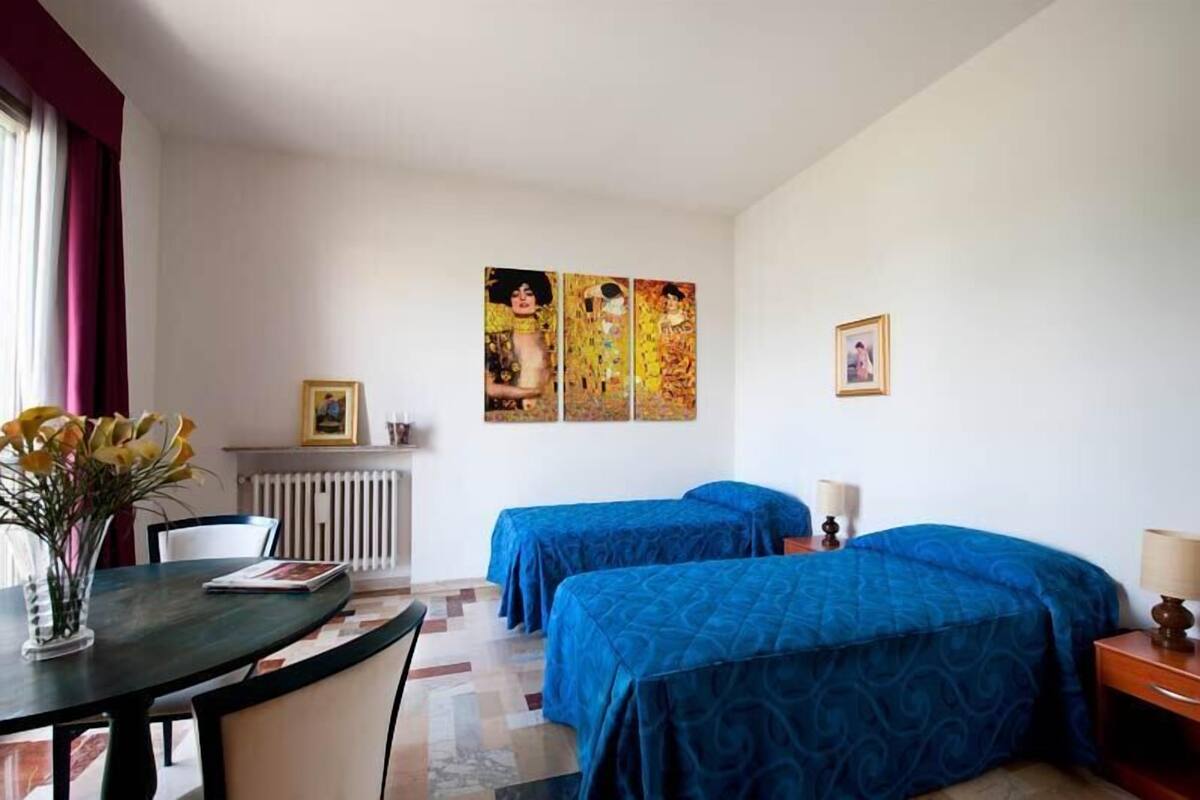 Una habitación de hotel con dos camas, una mesa con un florero y un cuadro en la pared.