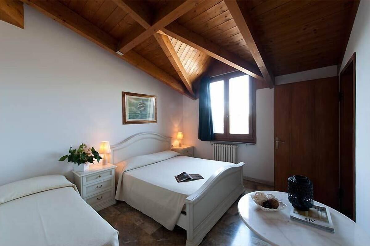 Un dormitorio con dos camas, techo de madera, ventana, radiador y mesita de noche con lámpara y un libro.