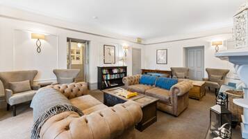 A spacious living room with a large sectional sofa, a coffee table, and a fireplace. There is a bookshelf and a door leading to another room.