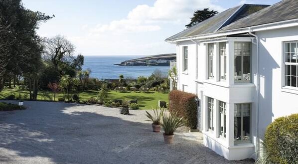 A white two-story house with a gravel driveway and a view of the sea. - The Rosevine (Truro)