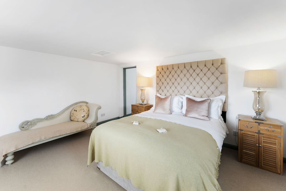 A hotel room with a large bed, a bedside table with a lamp, a wooden nightstand, and a bench with a cushion.