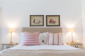 A neatly made bed with striped pillows, two framed sailboat pictures on the wall, and bedside lamps on nightstands. - The Rosevine (Truro)