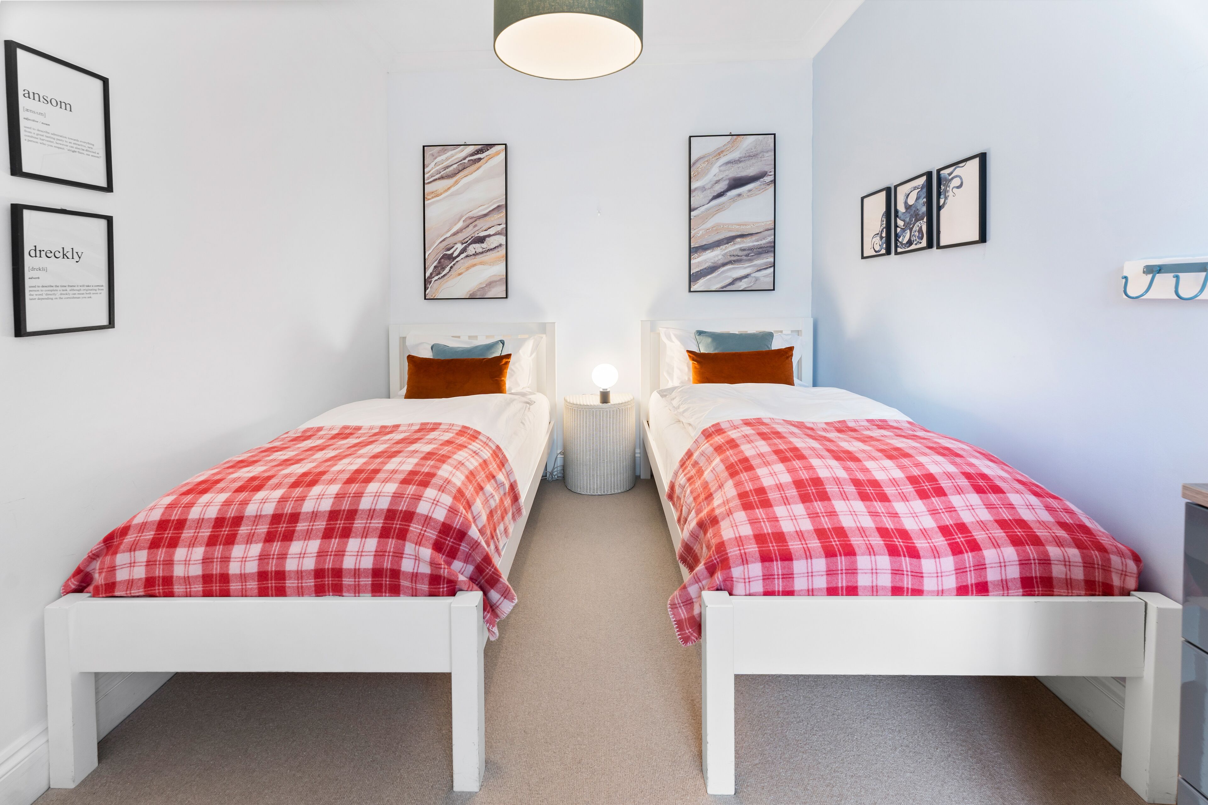 A room with two beds, each with a red plaid bedspread, white headboards, and a bedside table with a lamp. There are framed pictures on the walls and a coat rack to the right.