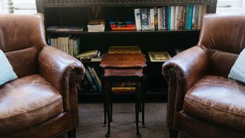 Zwei braune Ledersessel gegenüber, dazwischen ein kleiner Holztisch, dahinter eine Bücherwand und im Hintergrund ein Fenster.
