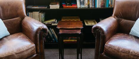 Two brown leather armchairs facing each other with a small wooden table in between, a bookshelf filled with books behind them, and a window in the background.