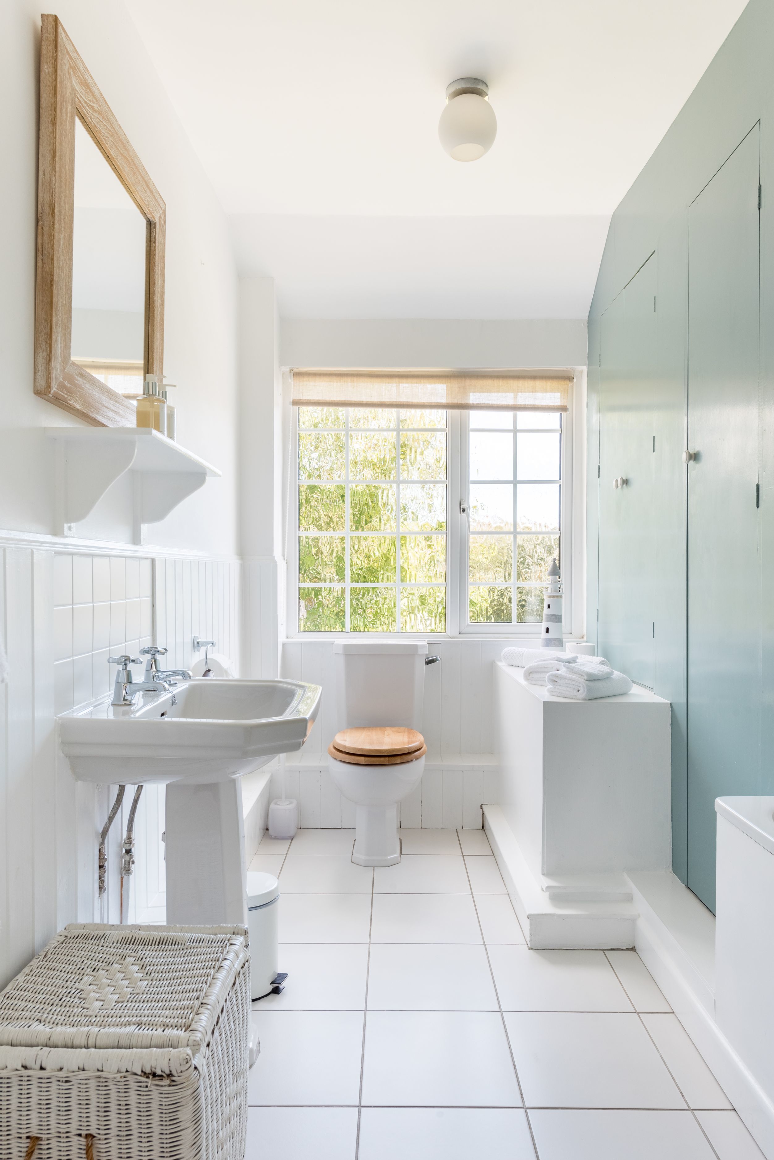 A bathroom with a white sink, toilet, and bathtub, a window with a view of greenery, and a wooden-framed mirror.