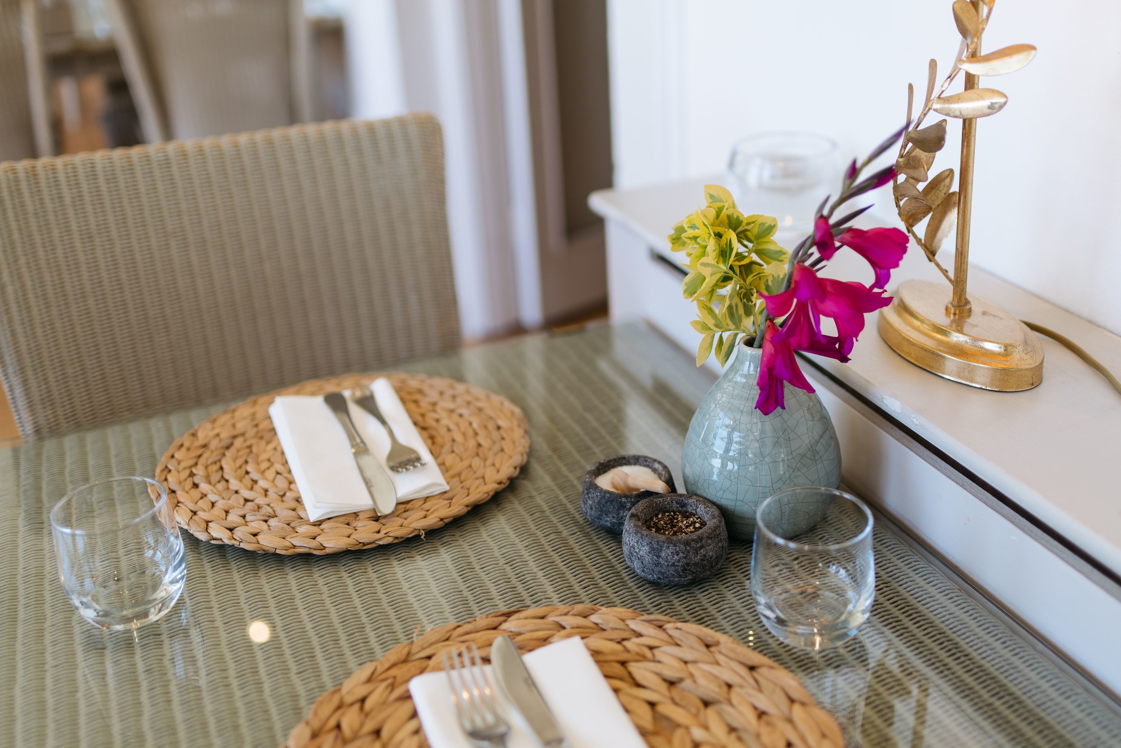 A dining table set with woven placemats, a vase with flowers, and a decorative gold stand.