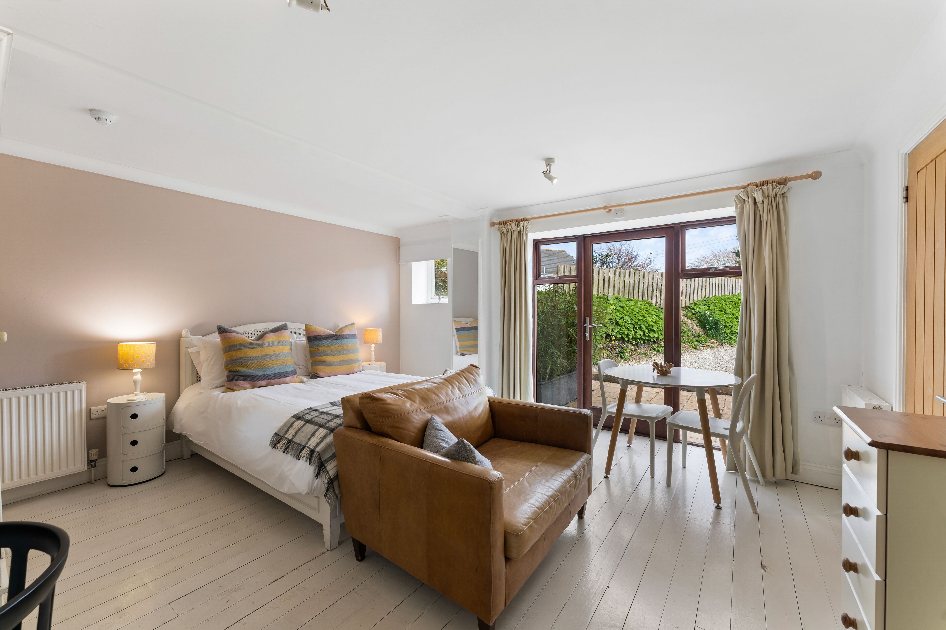 A bedroom with a bed, a brown leather sofa, a small table with chairs, and a view of the garden through a large window.