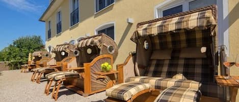 Outdoor seating with wooden chairs and a sofa, arranged in front of a building with large windows and a yellow facade.
