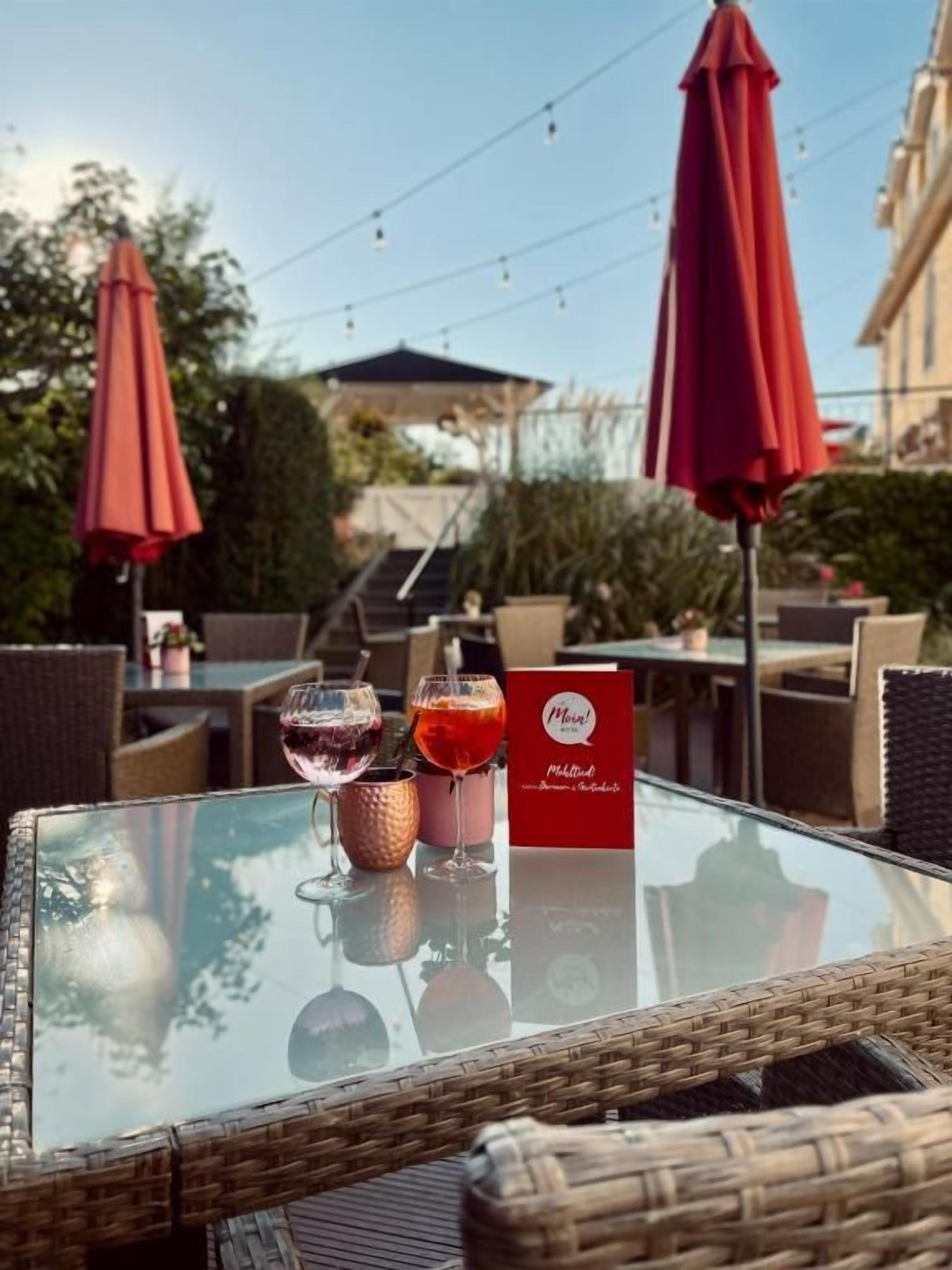 An outdoor dining area with wicker furniture, red umbrellas, and a glass table with a menu and two glasses of beverages.