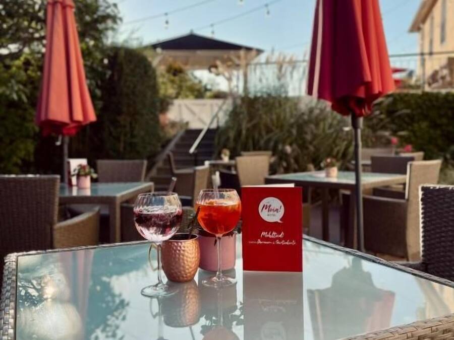 Un espacio de comedor al aire libre con mobiliario de mimbre, sombrillas rojas y una mesa de vidrio con un menú y dos vasos de bebidas.