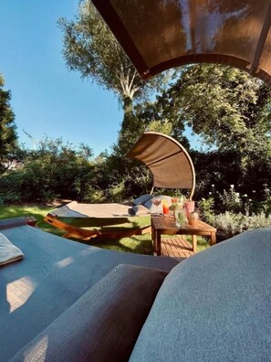 A hammock under a shade structure, a wooden table with drinks, and a grassy area with trees. - Moin! Hotel Cuxhaven (Cuxhaven)