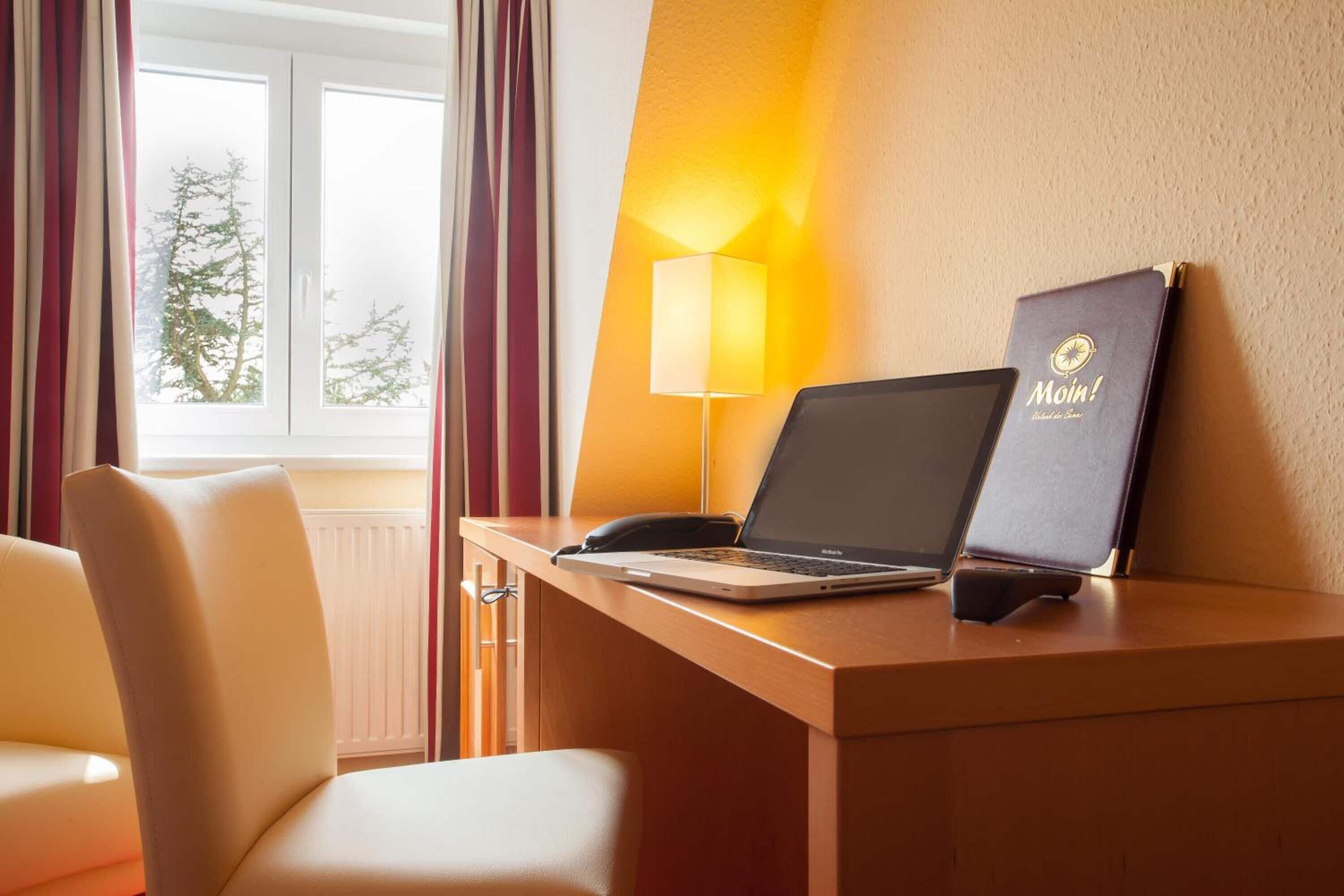 Hotellihuoneessa on työpöytä, kannettava tietokone, puhelin, lamppu ja seinällä roikkuu kirja, jonka otsikko on ”Moin!”.