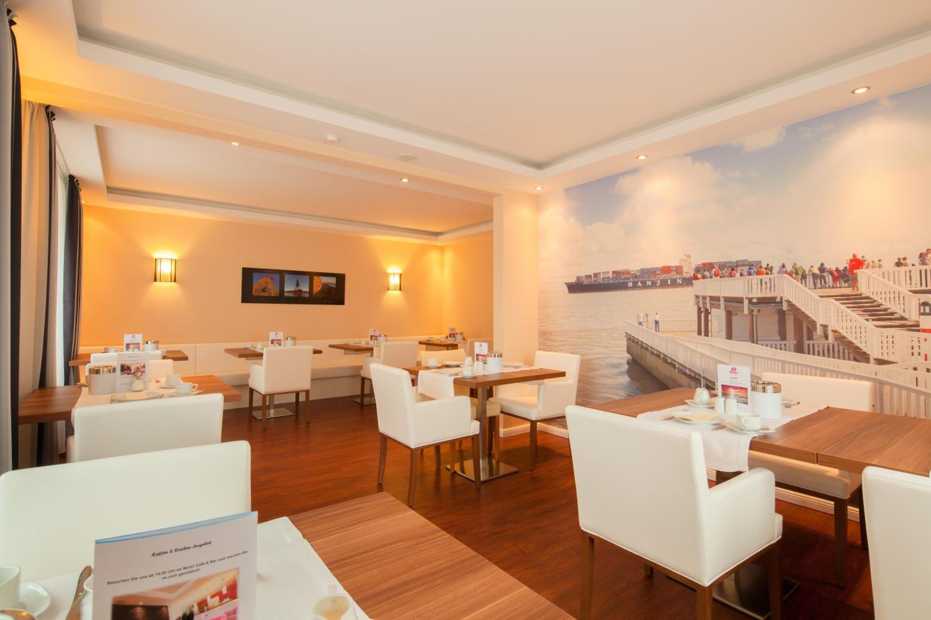 Un comedor de hotel con mesas y sillas, un mural de un puente y una escalera que conduce a un balcón.