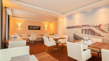 Un comedor de hotel con mesas y sillas, un mural de un puente y una escalera que conduce a un balcón.
