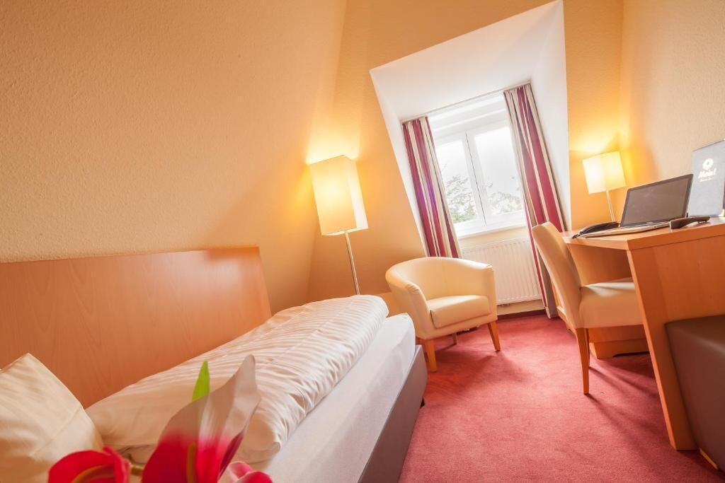 A hotel room with a bed, a desk with a laptop, a chair, and a window with curtains.