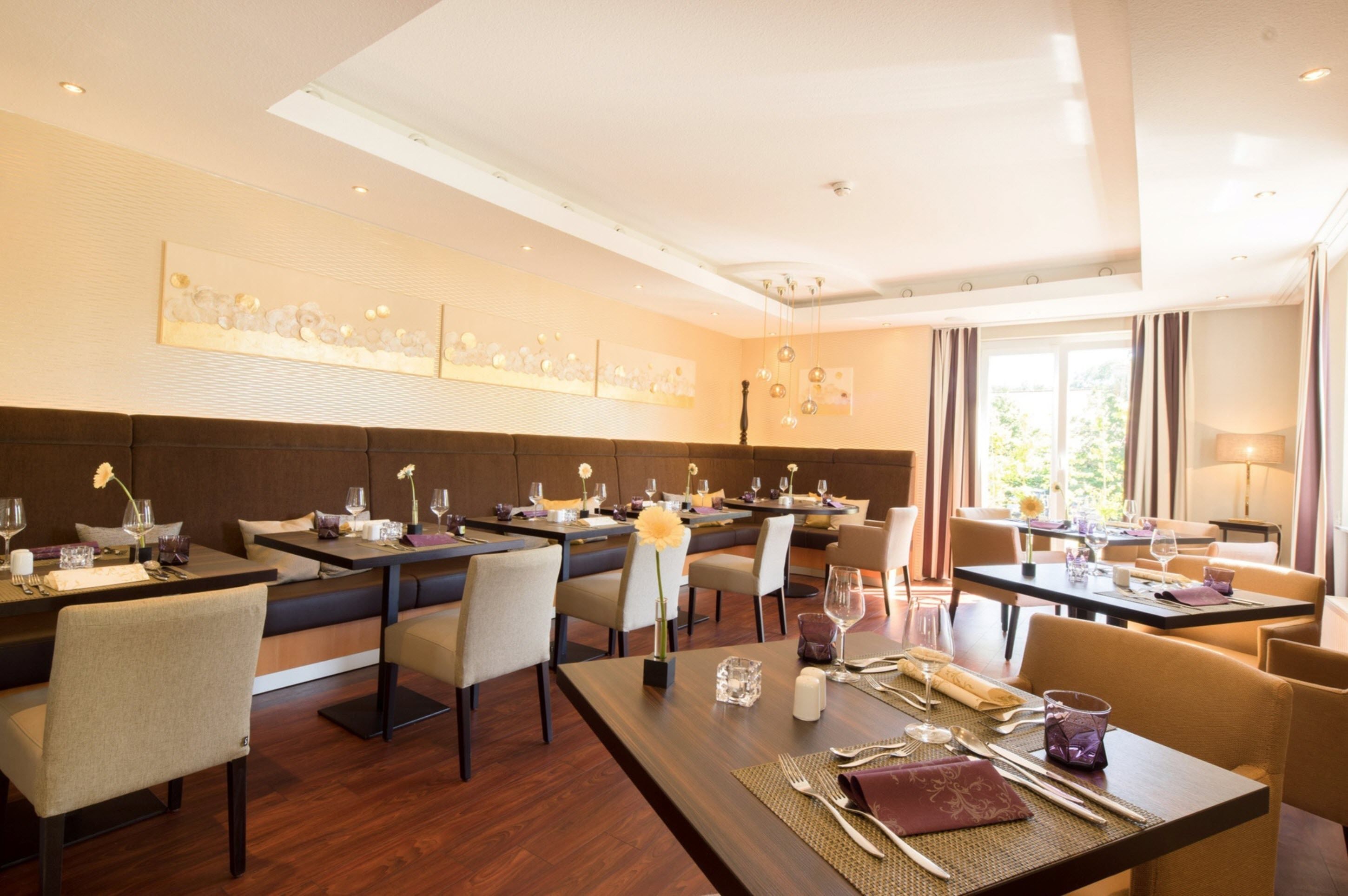 A modern dining area with multiple tables and chairs, a high ceiling with recessed lighting, and artwork on the walls.