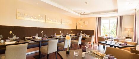 A modern dining area with multiple tables and chairs, a high ceiling with recessed lighting, and artwork on the walls.