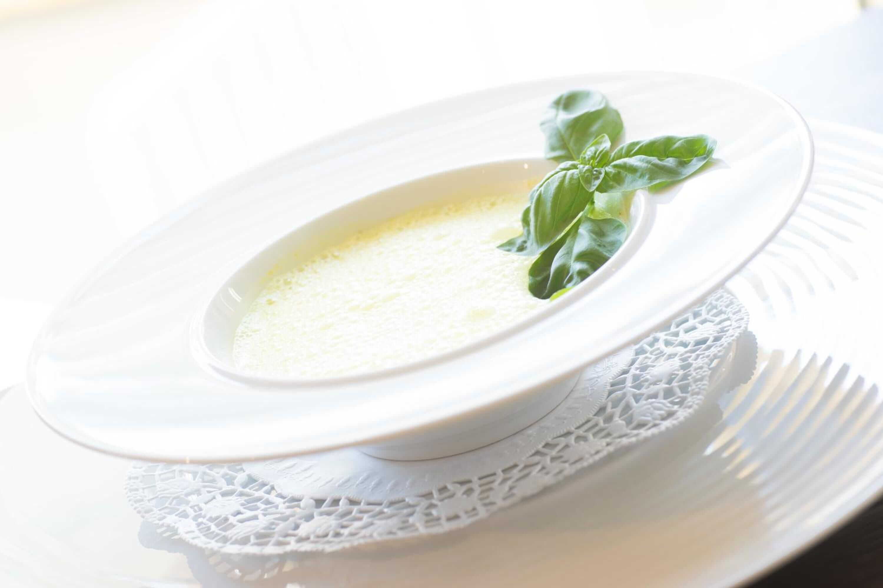 A white ceramic bowl with a creamy substance, garnished with basil leaves, placed on a white plate with a lace doily underneath.