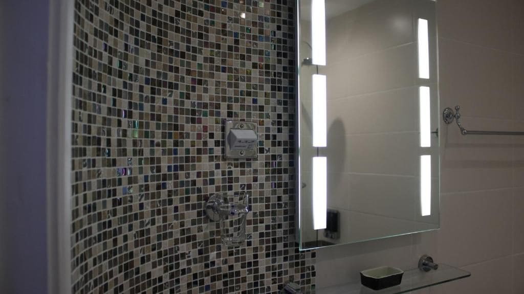 A bathroom with a mosaic tile wall, a glass shower enclosure, a mirror with a light, and a small sink with a soap dish.