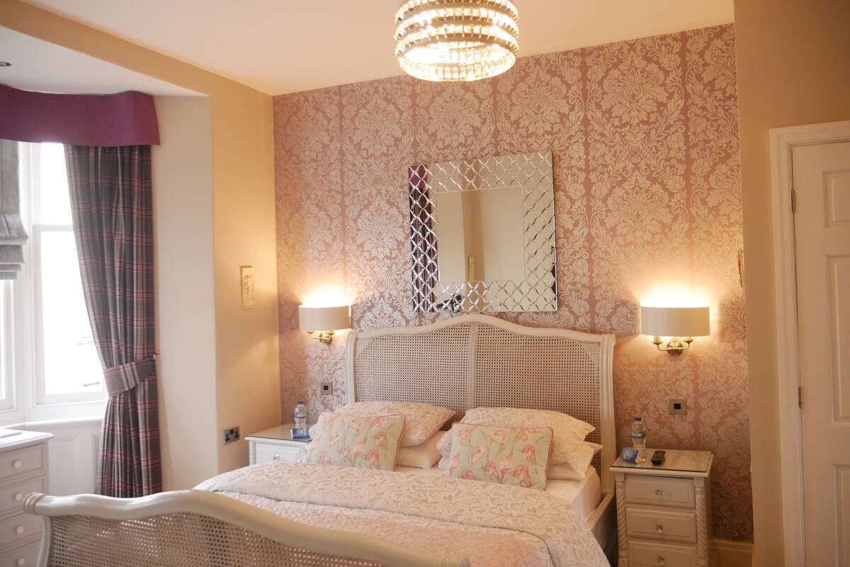 A bedroom with a bed, two bedside tables, a nightstand, a chandelier, and a mirror on the wall.