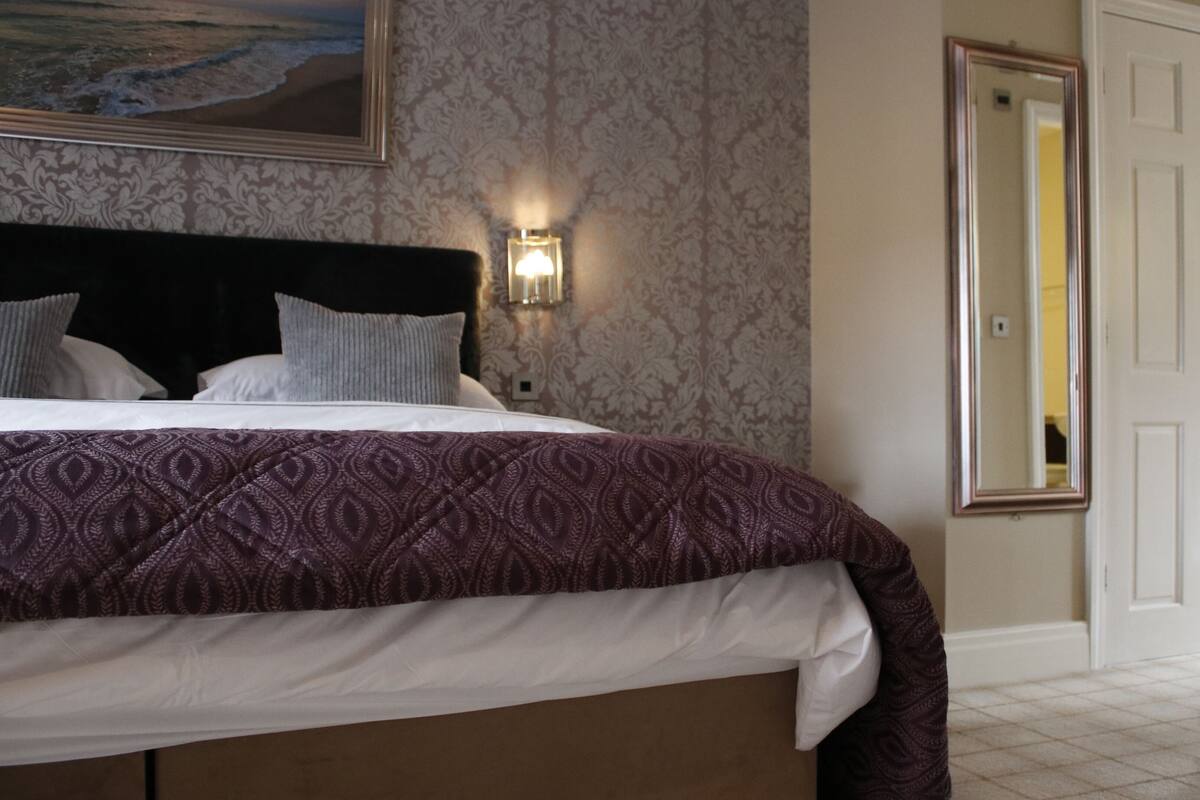 A neatly made bed with a patterned bedspread and pillows, a wall-mounted light fixture, and a door with a mirror.