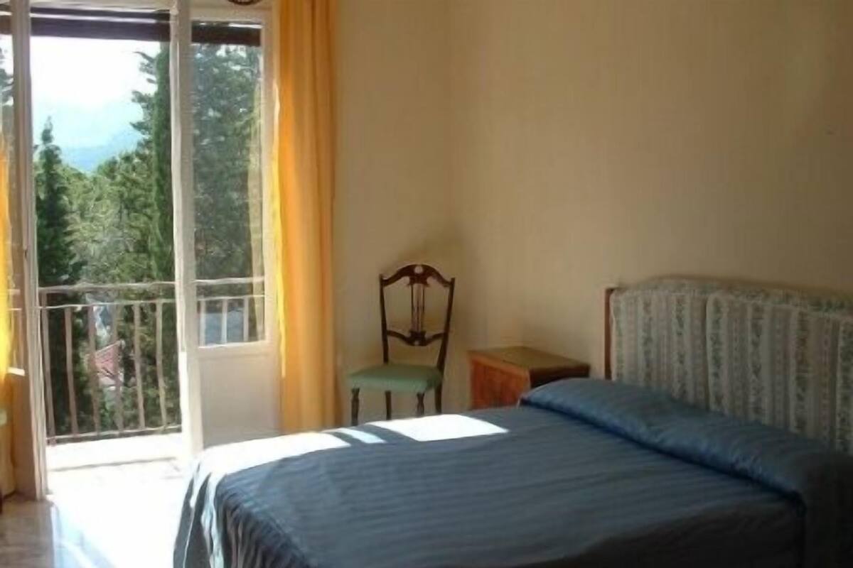 A bedroom with a bed, a chair, and a small table. There is a window with a view of trees and a balcony.