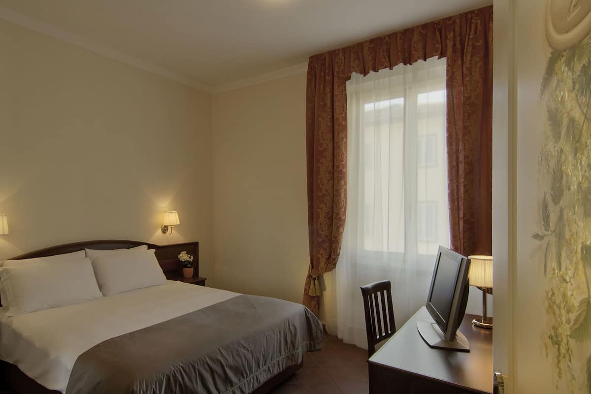 A hotel room with a bed, bedside lamps, a desk with a computer, and a window with curtains.