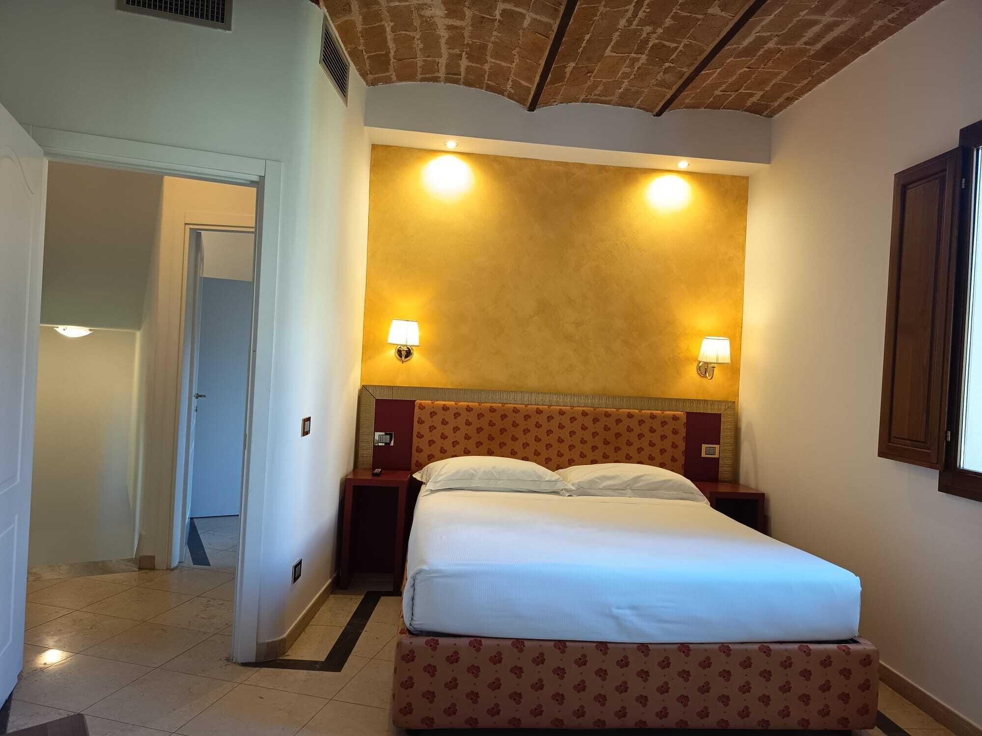 A hotel room with a bed, two bedside lamps, a headboard with a floral pattern, and a door leading to another room.